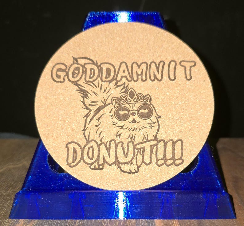 K&ouml;nnte beinhalten: Ein runder, beigefarbener Korkuntersetzer mit einer dunkelbraunen Lasergravur eines flauschigen Katzenmotivs, das eine Krone und eine runde Sonnenbrille tr&auml;gt. Der Text "GODDAMNIT DONUT!!!" ist in fetter Schrift &uuml;ber und unter der Katze eingraviert.