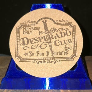 K&ouml;nnte beinhalten: Ein runder beigefarbener Korkuntersetzer mit dunkelbraunem Gravurdesign. Der Text lautet Members Only Desperado Club So Fun It Hurts! mit einer zentralen Dolchgrafik und einem kleinen Totenkopf-Symbol in einem dekorativen rechteckigen Rahmen.