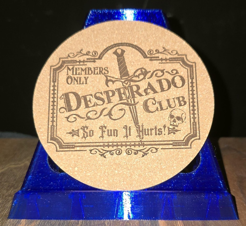 K&ouml;nnte beinhalten: Ein runder beigefarbener Korkuntersetzer mit dunkelbraunem Gravurdesign. Der Text lautet Members Only Desperado Club So Fun It Hurts! mit einer zentralen Dolchgrafik und einem kleinen Totenkopf-Symbol in einem dekorativen rechteckigen Rahmen.