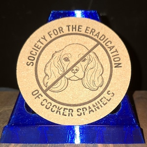 K&ouml;nnte beinhalten: Ein runder, beigefarbener Korkuntersetzer mit einer dunkelbraunen Strichzeichnung eines Cocker Spaniel-Gesichts, das von einer diagonalen Linie durchkreuzt wird. Der Text "SOCIETY FOR THE ERADICATION OF COCKER SPANIELS" steht kreisf&ouml;rmig darum.