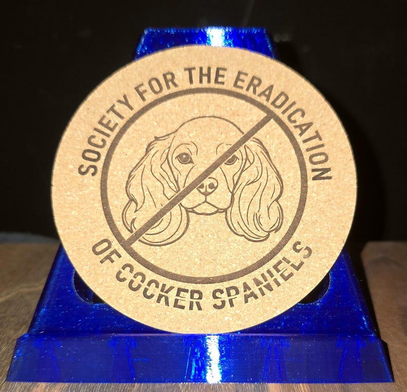 K&ouml;nnte beinhalten: Ein runder, beigefarbener Korkuntersetzer mit einer dunkelbraunen Strichzeichnung eines Cocker Spaniel-Gesichts, das von einer diagonalen Linie durchkreuzt wird. Der Text "SOCIETY FOR THE ERADICATION OF COCKER SPANIELS" steht kreisf&ouml;rmig darum.
