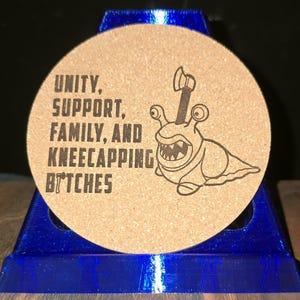 K&ouml;nnte beinhalten: Ein runder Untersetzer aus Kork mit einem schwarzen Aufdruck, der eine Schnecke mit einer Axt im Geh&auml;use zeigt. Der Text lautet: UNITY, SUPPORT, FAMILY, AND KNEECAPPING BITCHES. Der Untersetzer steht auf einem blauen Kunststoffst&auml;nder.