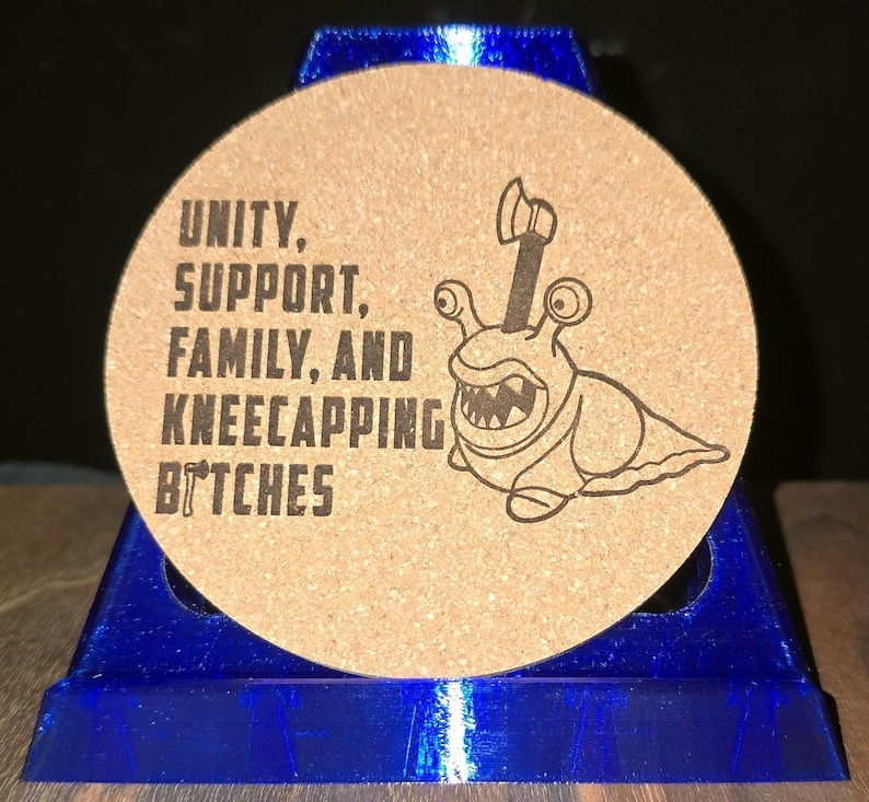 K&ouml;nnte beinhalten: Ein runder Untersetzer aus Kork mit einem schwarzen Aufdruck, der eine Schnecke mit einer Axt im Geh&auml;use zeigt. Der Text lautet: UNITY, SUPPORT, FAMILY, AND KNEECAPPING BITCHES. Der Untersetzer steht auf einem blauen Kunststoffst&auml;nder.