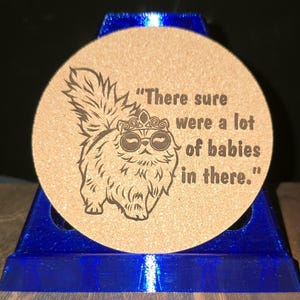 K&ouml;nnte beinhalten: Ein runder, hellbrauner Korkuntersetzer mit einer dunkelbraunen, lasergravierten Illustration einer flauschigen Katze, die eine Krone und eine runde Brille tr&auml;gt. Der Text "There sure were a lot of babies in there." steht rechts daneben.