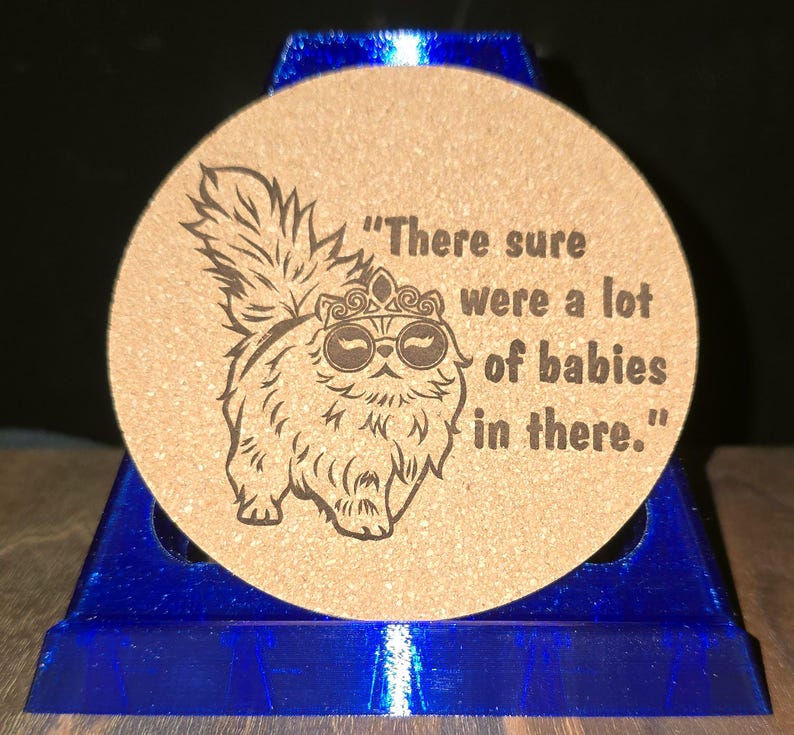 K&ouml;nnte beinhalten: Ein runder, hellbrauner Korkuntersetzer mit einer dunkelbraunen, lasergravierten Illustration einer flauschigen Katze, die eine Krone und eine runde Brille tr&auml;gt. Der Text "There sure were a lot of babies in there." steht rechts daneben.