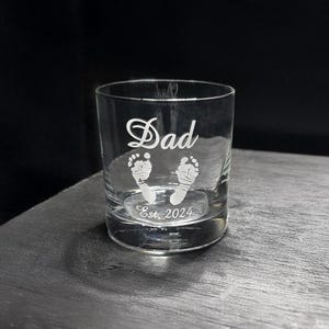 Może przedstawiać: Przezroczysty szklany kubek z napisem "Dad" w ozdobnym kroju pisma, z dwoma wzorami stópek dziecka i tekstem "Est. 2024". Potencjalny prezent dla nowego taty.