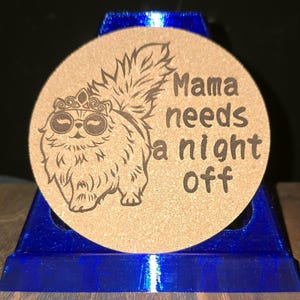 K&ouml;nnte beinhalten: Ein runder, beigefarbener Korkuntersetzer mit einer lasergravierten Illustration einer flauschigen Katze, die eine Krone und eine runde Sonnenbrille tr&auml;gt, sowie dem Text "Mama needs a night off" in einer dunklen, fetten, serifenlosen Schrift.
