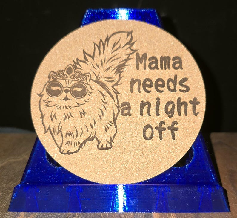 K&ouml;nnte beinhalten: Ein runder, beigefarbener Korkuntersetzer mit einer lasergravierten Illustration einer flauschigen Katze, die eine Krone und eine runde Sonnenbrille tr&auml;gt, sowie dem Text "Mama needs a night off" in einer dunklen, fetten, serifenlosen Schrift.
