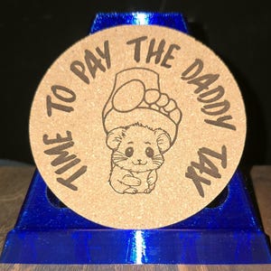 K&ouml;nnte beinhalten: Ein runder Untersetzer aus beigem Kork mit einer dunkelbraunen, lasergravierten Illustration eines kleinen Hamsters, &uuml;ber dessen Kopf sich ein menschlicher Fu&szlig; befindet. Der Text "TIME TO PAY THE DADDY TAX" ist kreisf&ouml;rmig eingraviert.