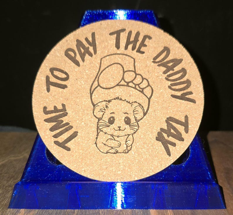 K&ouml;nnte beinhalten: Ein runder Untersetzer aus beigem Kork mit einer dunkelbraunen, lasergravierten Illustration eines kleinen Hamsters, &uuml;ber dessen Kopf sich ein menschlicher Fu&szlig; befindet. Der Text "TIME TO PAY THE DADDY TAX" ist kreisf&ouml;rmig eingraviert.