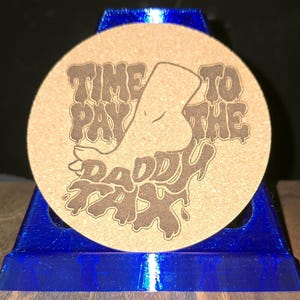 K&ouml;nnte beinhalten: Ein runder, hellbrauner Korkuntersetzer mit einem dunkelbraunen, lasergravierten Motiv eines Fu&szlig;es und dem Text "TIME TO PAY THE DADDY TAX" in einer stilisierten, tropfenden Schriftart. Der Untersetzer steht auf einem leuchtend blauen, eckigen Kunststoffst&auml;nder.