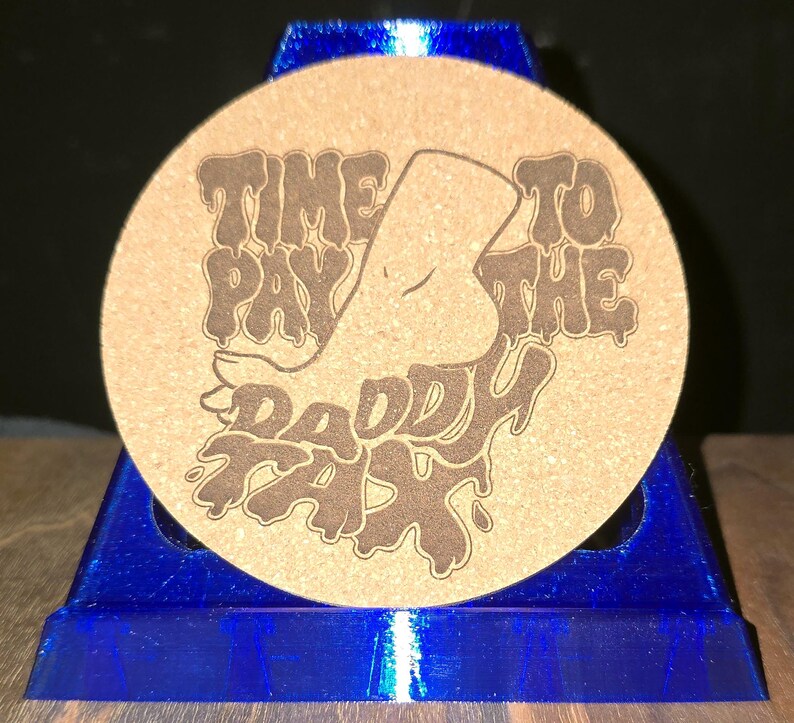 K&ouml;nnte beinhalten: Ein runder, hellbrauner Korkuntersetzer mit einem dunkelbraunen, lasergravierten Motiv eines Fu&szlig;es und dem Text "TIME TO PAY THE DADDY TAX" in einer stilisierten, tropfenden Schriftart. Der Untersetzer steht auf einem leuchtend blauen, eckigen Kunststoffst&auml;nder.
