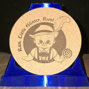 K&ouml;nnte beinhalten: Ein runder, beigefarbener Korkuntersetzer mit einer dunkelbraunen Lasergravur eines Charakters, der einen Hut mit Feder und eine Lederhose tr&auml;gt und einen Lutscher h&auml;lt. Der Text "Run, Little G&uuml;nter, Run!" ist &uuml;ber dem Kopf abgebildet.