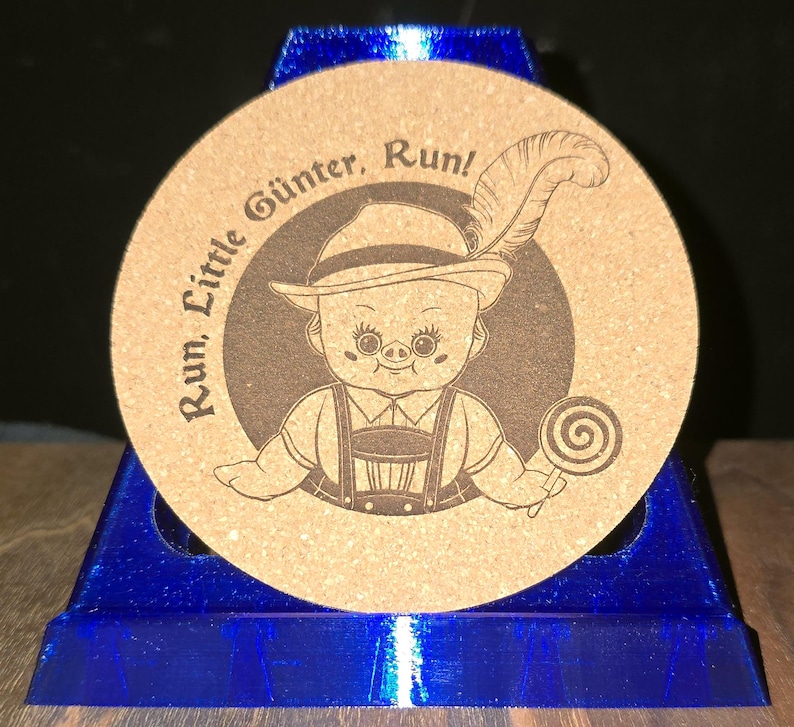 K&ouml;nnte beinhalten: Ein runder, beigefarbener Korkuntersetzer mit einer dunkelbraunen Lasergravur eines Charakters, der einen Hut mit Feder und eine Lederhose tr&auml;gt und einen Lutscher h&auml;lt. Der Text "Run, Little G&uuml;nter, Run!" ist &uuml;ber dem Kopf abgebildet.
