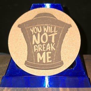 K&ouml;nnte beinhalten: Ein runder, beigefarbener Korkuntersetzer mit einer dunkelbraunen Lasergravur, die eine &auml;rmellose Jeansweste darstellt. Der Text "YOU WILL NOT BREAK ME" ist mittig im Westendesign platziert und in einer fetten, handgeschriebenen Schriftart.