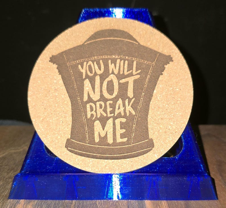 K&ouml;nnte beinhalten: Ein runder, beigefarbener Korkuntersetzer mit einer dunkelbraunen Lasergravur, die eine &auml;rmellose Jeansweste darstellt. Der Text "YOU WILL NOT BREAK ME" ist mittig im Westendesign platziert und in einer fetten, handgeschriebenen Schriftart.