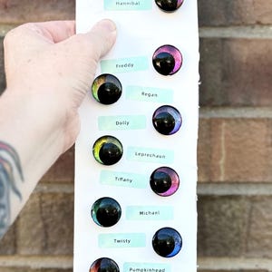 May include: A selection of colorful, iridescent glass eyes with black pupils, each paired with a name tag. Names include "Jason", "Chucky", "Hannibal", "Freddy", "Regan", "Dolly", "Leprechaun", "Tiffany", "Michael", "Twisty", "Pumpkinhead", "Dancing clown", and "Jane Doe".