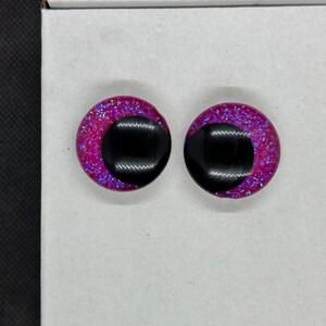 May include: A pair of circular stud earrings featuring a vibrant magenta base infused with fine blue glitter. Each earring has a large, smooth, black circular inlay positioned off-center, creating a distinct contrast against the sparkling pink background.