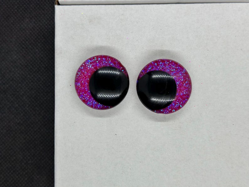 May include: A pair of circular stud earrings featuring a vibrant magenta base infused with fine blue glitter. Each earring has a large, smooth, black circular inlay positioned off-center, creating a distinct contrast against the sparkling pink background.