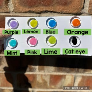 May include: A white rectangular display card featuring eight circular cutouts, each containing a colored cabochon. The colors are labeled with green text: Purple, Lemon, Blue, Orange, Mint, Pink, Lime, and Cat eye. The cabochons are translucent.