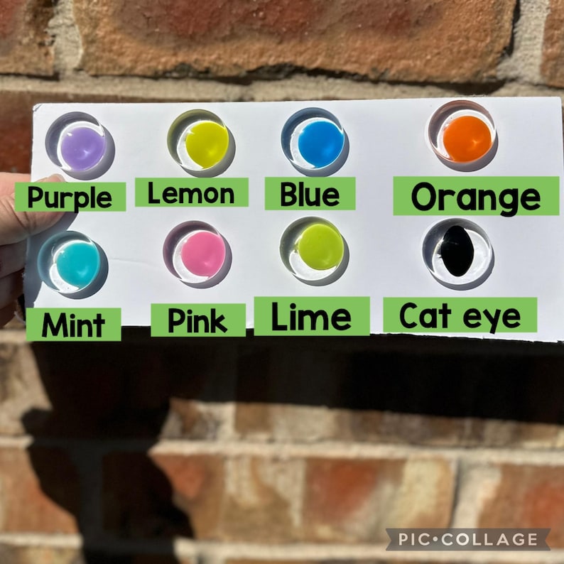 May include: A white rectangular display card featuring eight circular cutouts, each containing a colored cabochon. The colors are labeled with green text: Purple, Lemon, Blue, Orange, Mint, Pink, Lime, and Cat eye. The cabochons are translucent.