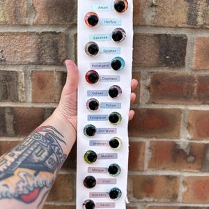 May include: A vertical white display card featuring fourteen pairs of round, glittery cabochon eyes in various colors, each labeled with names including White Noise, Arson, Hellfire, Banshee, Splinter, Poltergeist, Countess, Cursed, Potion, Ectoplasm, Still Water, Skeleton, Monster, Morgue, Crypt, Boo-berry, and Dexter.