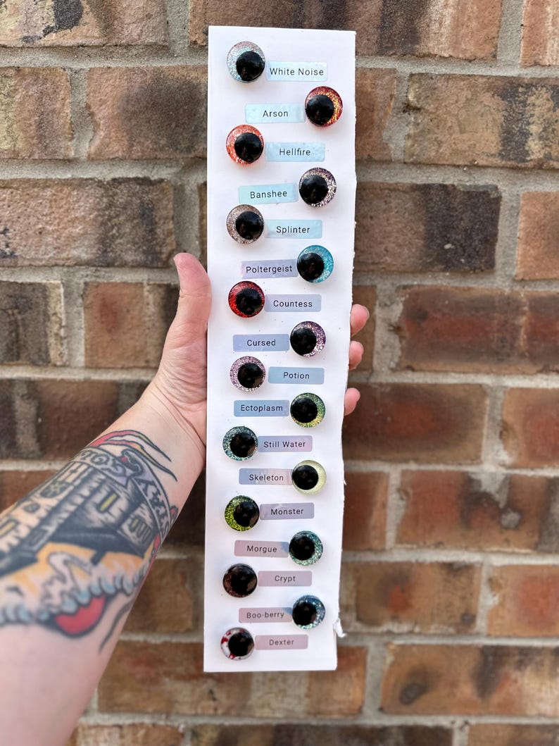 May include: A vertical white display card featuring fourteen pairs of round, glittery cabochon eyes in various colors, each labeled with names including White Noise, Arson, Hellfire, Banshee, Splinter, Poltergeist, Countess, Cursed, Potion, Ectoplasm, Still Water, Skeleton, Monster, Morgue, Crypt, Boo-berry, and Dexter.