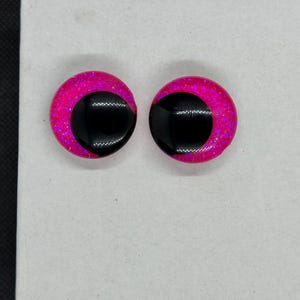 May include: A pair of circular craft eyes featuring a vibrant hot pink glitter base with a large, solid black pupil offset to one side. These round, smooth-surfaced components have a glossy finish and are designed for use in various creative projects.
