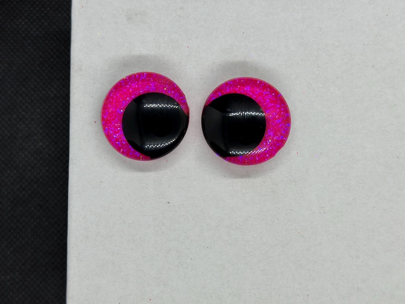 May include: A pair of circular craft eyes featuring a vibrant hot pink glitter base with a large, solid black pupil offset to one side. These round, smooth-surfaced components have a glossy finish and are designed for use in various creative projects.