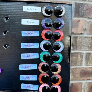 May include: A black display board featuring eight pairs of circular craft eyes with glittery irises in silver, purple, red, light blue, green, pink, orange, and light pink. Each pair is labeled with text: Coffin Nail, Hexing, Lobotomy, Frost Bite, Goblin, Brains, Patch, and Salem.