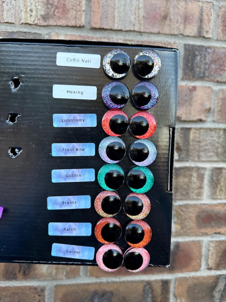 May include: A black display board featuring eight pairs of circular craft eyes with glittery irises in silver, purple, red, light blue, green, pink, orange, and light pink. Each pair is labeled with text: Coffin Nail, Hexing, Lobotomy, Frost Bite, Goblin, Brains, Patch, and Salem.