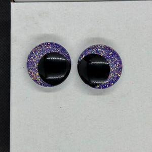May include: A pair of circular stud earrings featuring a glossy black crescent shape set against a background of deep purple resin embedded with fine, multi-colored iridescent glitter particles that catch the light across the surface.