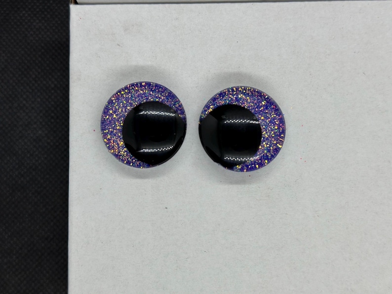 May include: A pair of circular stud earrings featuring a glossy black crescent shape set against a background of deep purple resin embedded with fine, multi-colored iridescent glitter particles that catch the light across the surface.