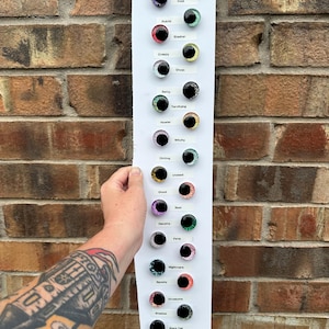 May include: A white vertical display board featuring multiple pairs of round, multi-colored glass cabochon eyes. Each pair is labeled with text including Naughty, Horrifying, Omen, Void, Putrid, Slasher, Creepy, Ghost, Batty, Terrifying, Howler, Witchy, Chilling, Undead, Ghoul, Boot, Decomp, Fang, Nightmare, Spooky, Gruesome, Shadow, Black Cat, and Frowzie.