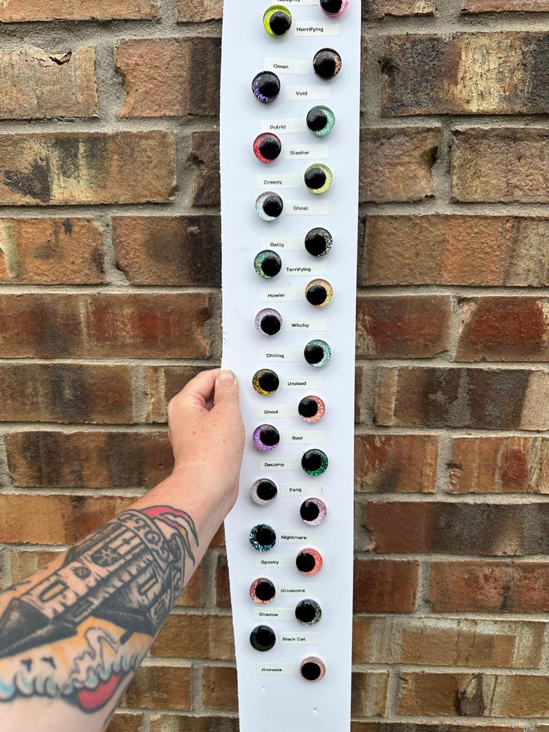 May include: A white vertical display board featuring multiple pairs of round, multi-colored glass cabochon eyes. Each pair is labeled with text including Naughty, Horrifying, Omen, Void, Putrid, Slasher, Creepy, Ghost, Batty, Terrifying, Howler, Witchy, Chilling, Undead, Ghoul, Boot, Decomp, Fang, Nightmare, Spooky, Gruesome, Shadow, Black Cat, and Frowzie.