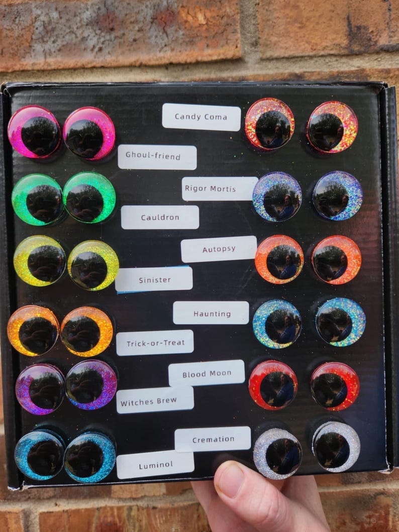May include: A black display box containing fourteen pairs of circular, glittery cabochon eyes with black pupils. The labels read: Candy Coma, Ghoul-friend, Rigor Mortis, Cauldron, Autopsy, Sinister, Haunting, Trick-or-Treat, Blood Moon, Witches Brew, and Luminal.