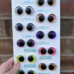 May include: A collection of round, glittery cabochon eyes arranged in pairs on a white display card. Each pair features a large black pupil and is labeled with names including Sarcophagus, Scythe, Ghastly, Twilight, Wicked, Spellbound, Love Spell, Toxic Waste, Mortified, and Zombie.