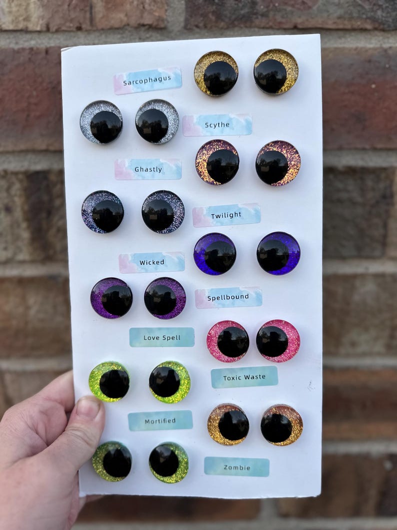 May include: A collection of round, glittery cabochon eyes arranged in pairs on a white display card. Each pair features a large black pupil and is labeled with names including Sarcophagus, Scythe, Ghastly, Twilight, Wicked, Spellbound, Love Spell, Toxic Waste, Mortified, and Zombie.