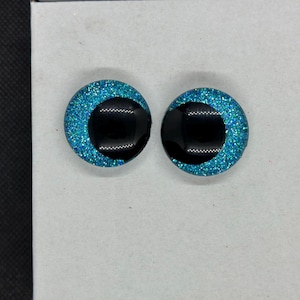 May include: A pair of circular cabochons featuring a bright turquoise glitter background with a large, solid black pupil centered in each. These round, eye-like pieces have a smooth, glossy finish and a flat back, designed for craft projects.
