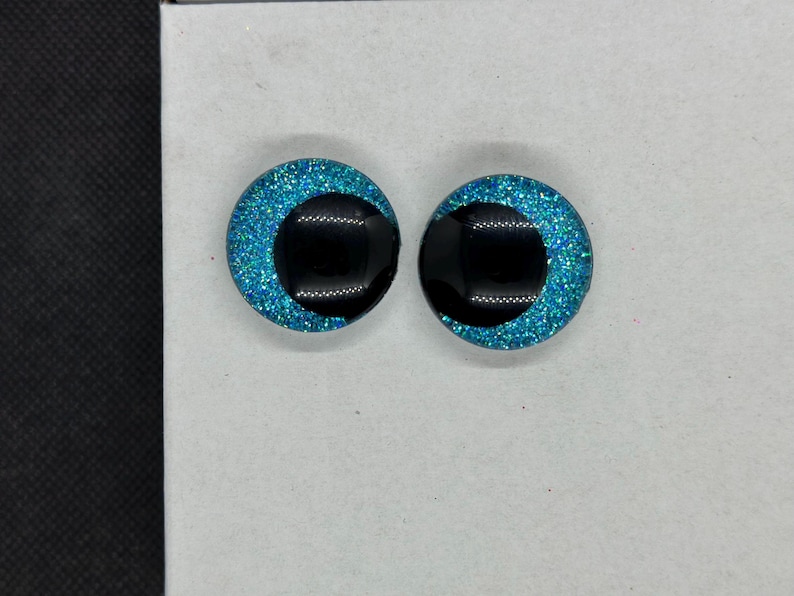 May include: A pair of circular cabochons featuring a bright turquoise glitter background with a large, solid black pupil centered in each. These round, eye-like pieces have a smooth, glossy finish and a flat back, designed for craft projects.