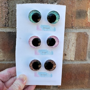 May include: Three pairs of circular cabochon eyes on a white card, labeled Dewey, Cujo, and Gizmo. The top pair is teal glitter, the middle pair is pink glitter, and the bottom pair is copper glitter. Each eye features a large, solid black pupil.