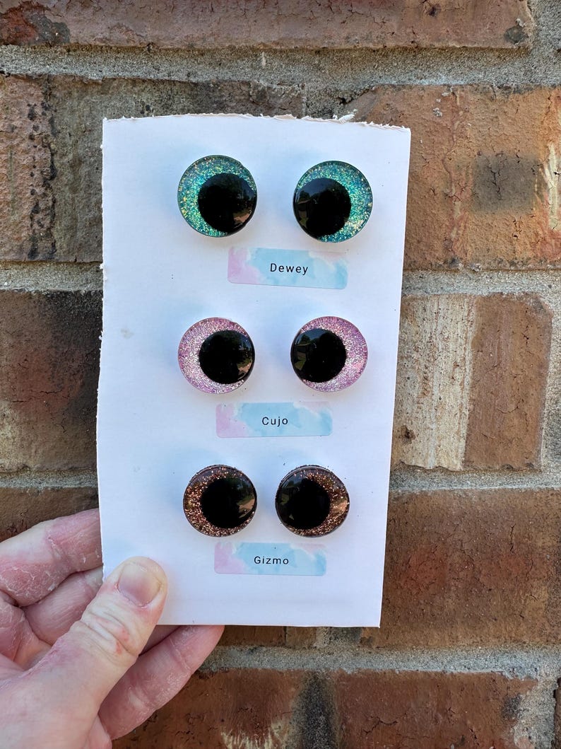 May include: Three pairs of circular cabochon eyes on a white card, labeled Dewey, Cujo, and Gizmo. The top pair is teal glitter, the middle pair is pink glitter, and the bottom pair is copper glitter. Each eye features a large, solid black pupil.