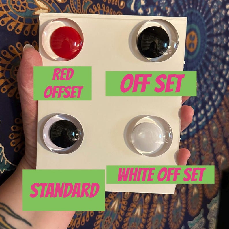 May include: A white rectangular card featuring four circular googly eyes. The top left eye has a red pupil, the top right has a black pupil, the bottom left has a black pupil, and the bottom right has a white pupil. Text labels include RED OFFSET, OFF SET, STANDARD, and WHITE OFF SET.