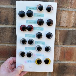 May include: A white display board featuring seven pairs of circular cabochon eyes with black pupils and various colored irises. The labels include Macabre, Reaper, Graveyard Dirt, Vampire, Casket, Tombstone, Gargoyle, Phantom, and Eerie, arranged in rows.
