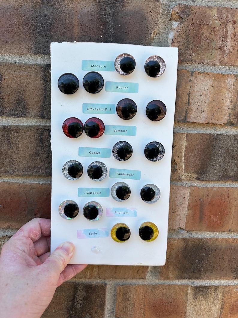 May include: A white display board featuring seven pairs of circular cabochon eyes with black pupils and various colored irises. The labels include Macabre, Reaper, Graveyard Dirt, Vampire, Casket, Tombstone, Gargoyle, Phantom, and Eerie, arranged in rows.