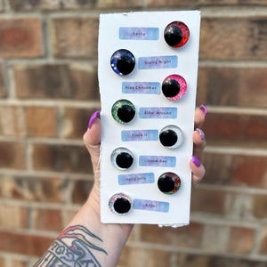 May include: A white card with eight different Christmas-themed googly eyes. The eyes are decorated with glitter and have different colors and patterns. The card has the names of each eye written below them: Santa, Starry Night, Pink Christmas, Elfin' Around, Flock It, Holly Jolly, Snow Day, and Angel.
