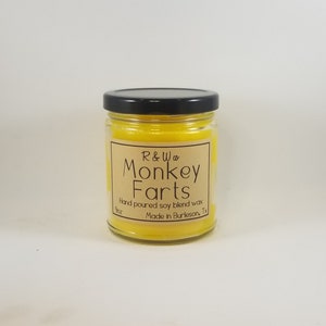 May include: A yellow glass jar candle with a black lid. The label reads "R & W co Monkey Farts" and "Hand poured soy blend wax Made in Burleson, Tx".