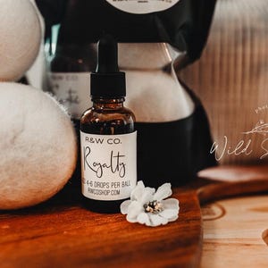 Puede incluir: Una botella de vidrio marrón con un gotero y una etiqueta blanca que dice "R&W CO. Royalty 4-6 Drops Per Ball RWCShop.com". La botella está sobre una superficie de madera con bolas de secado de lana blanca.