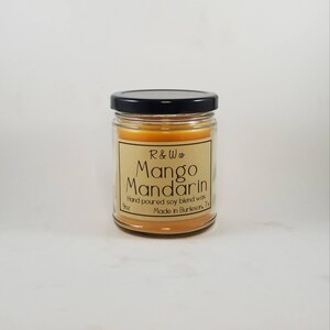 May include: A 9oz candle in a glass jar with a black lid. The candle is a light orange colour and has a label that says "R&W co Mango Mandarin Hand poured soy blend wax Made in Burleson, Tx".