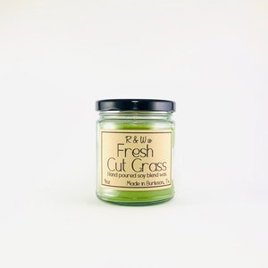 May include: A glass jar candle with a black lid. The label is brown with white text that reads "R&W co Fresh Cut Grass Hand poured soy blend wax 9oz Made in Burleson, Tx".